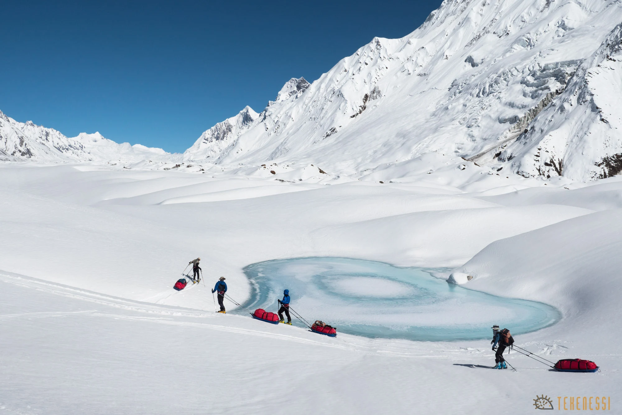 Skiers pulling sleds across a snowy Karakoram glacier beside a turquoise meltwater pool